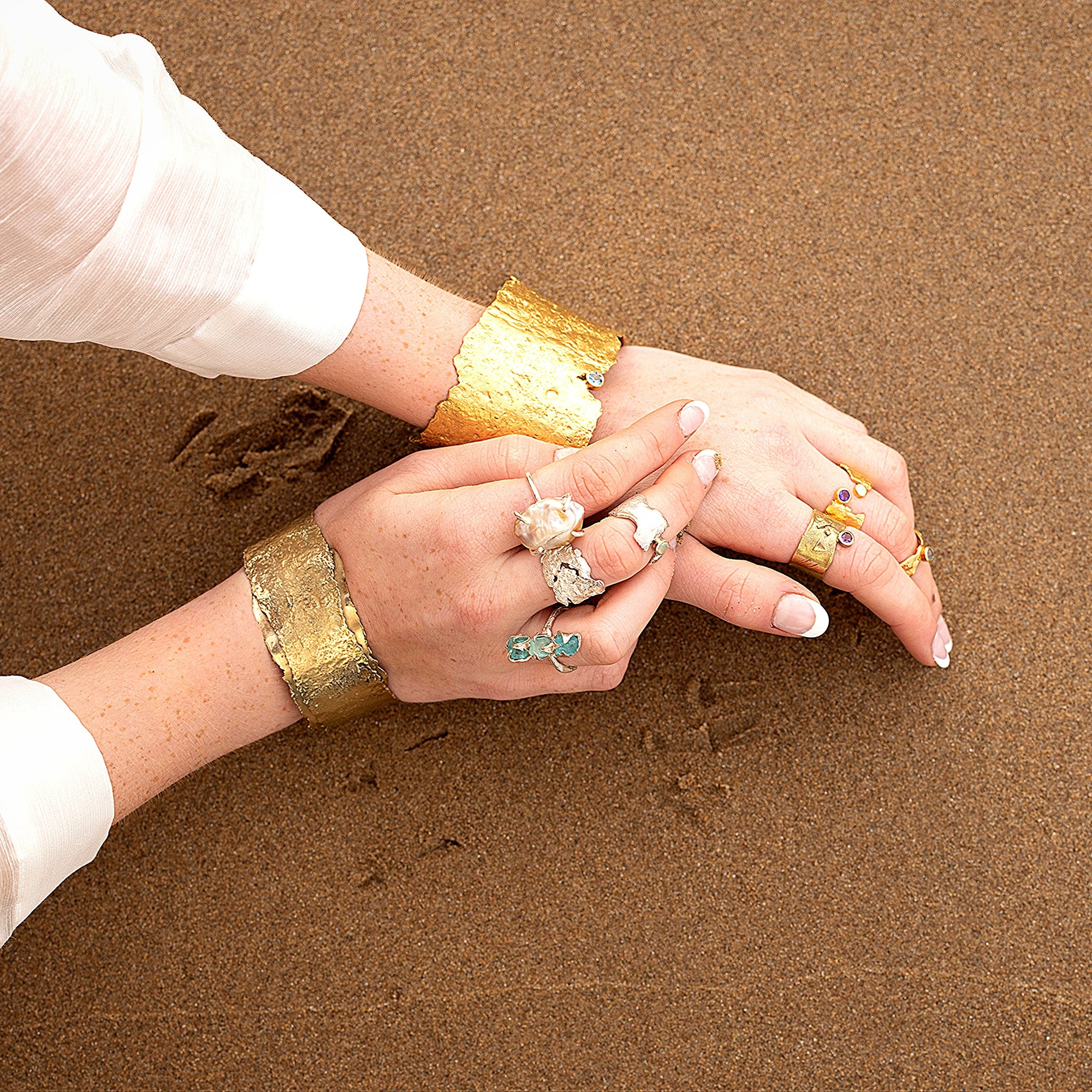 Close-up of hands with boho irish gold cuff bangles and unique gemstones rings on a sandy beach in Ireland